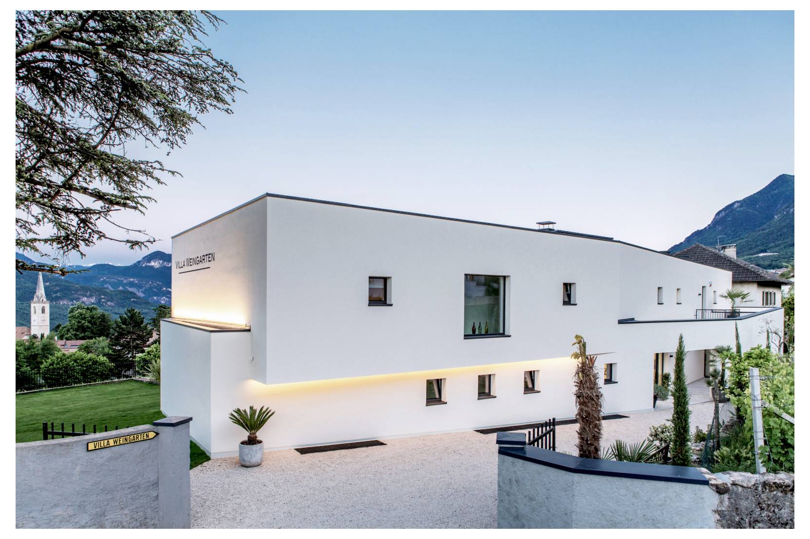 Bâtiment blanc moderne et minimaliste à l'extérieur éclairé au crépuscule, implanté sur une allée en gravier, avec des montagnes et des arbres en arrière-plan.