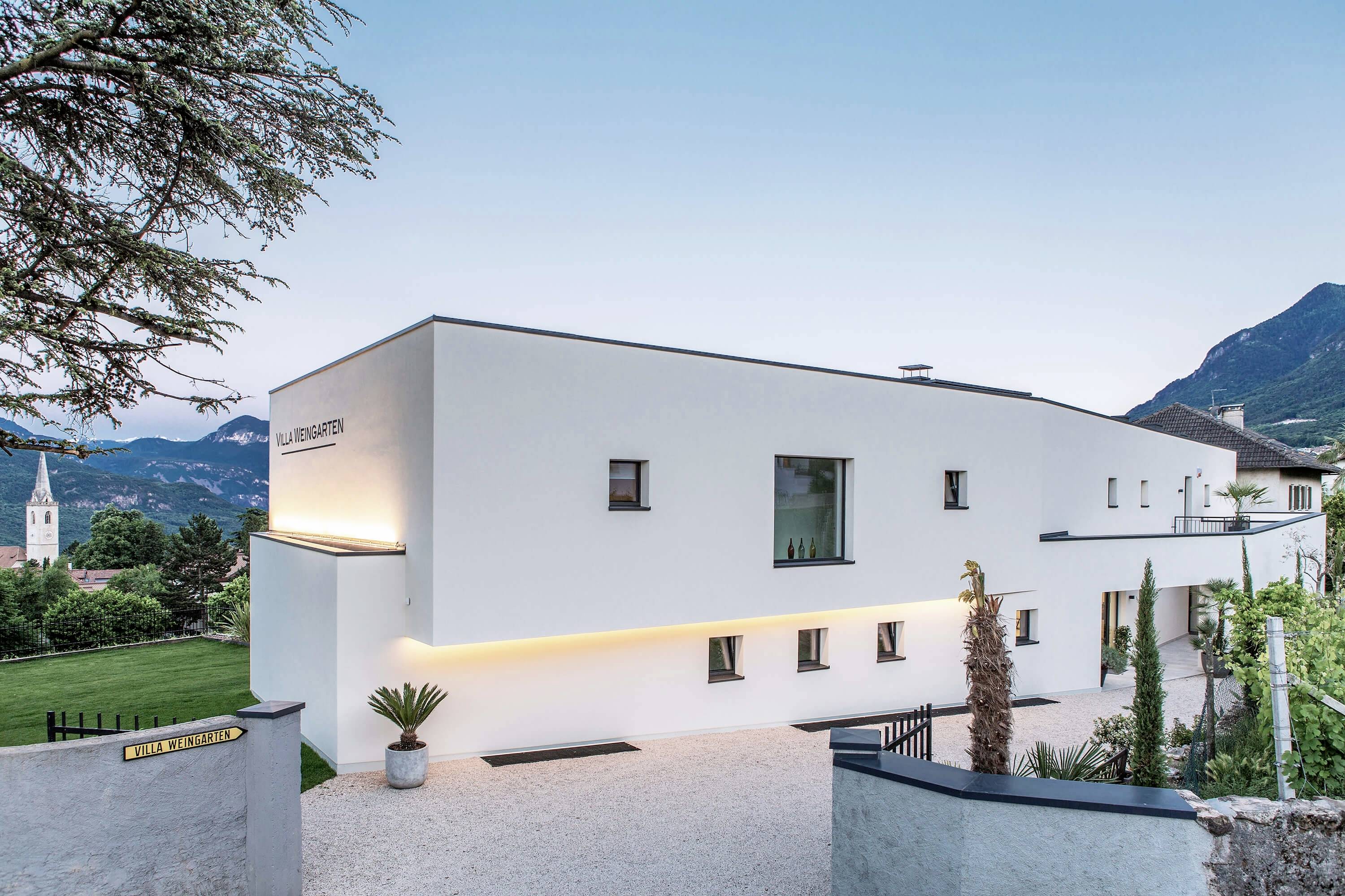 Un bâtiment blanc moderne aux lignes épurées, entouré de montagnes et d'espaces verts, en plein jour. Une église est visible au loin sur la gauche.