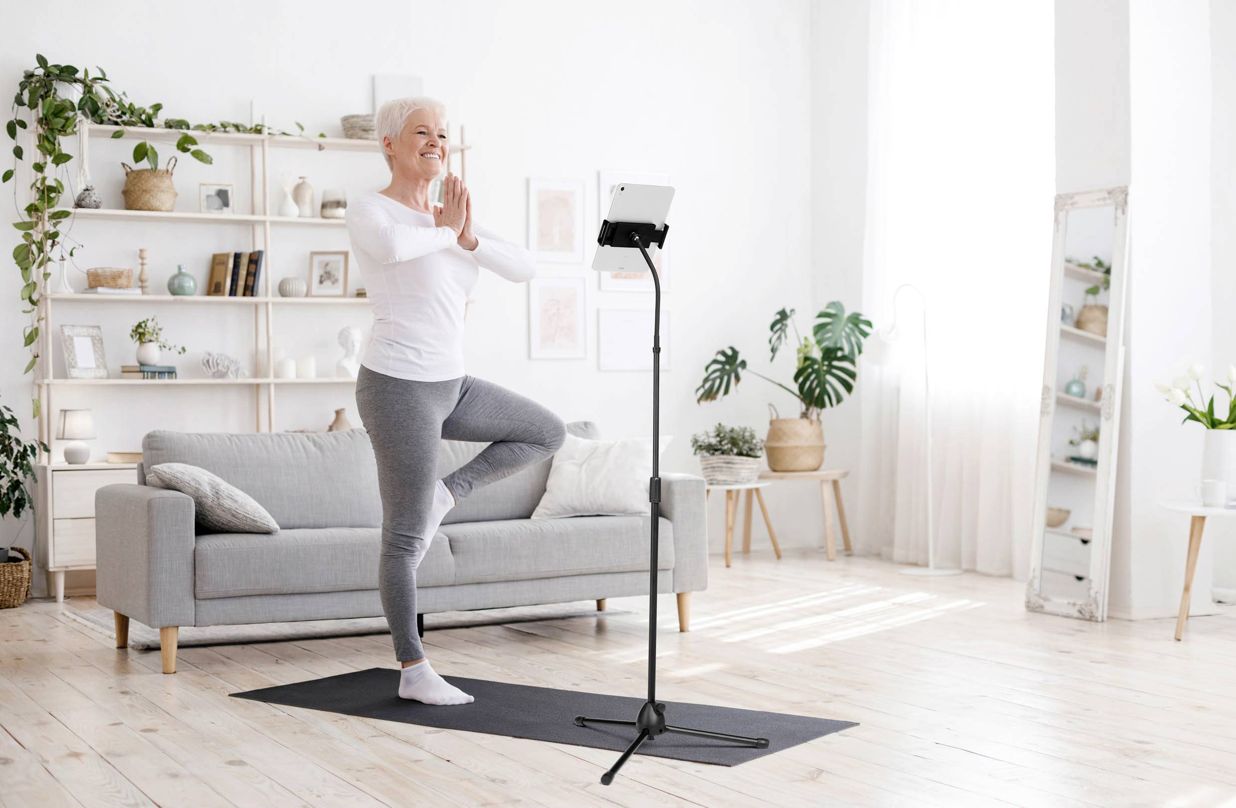 Une personne âgée pratique le yoga dans un salon lumineux. Elle est debout sur un tapis en position d'arbre devant une tablette posée sur un support.