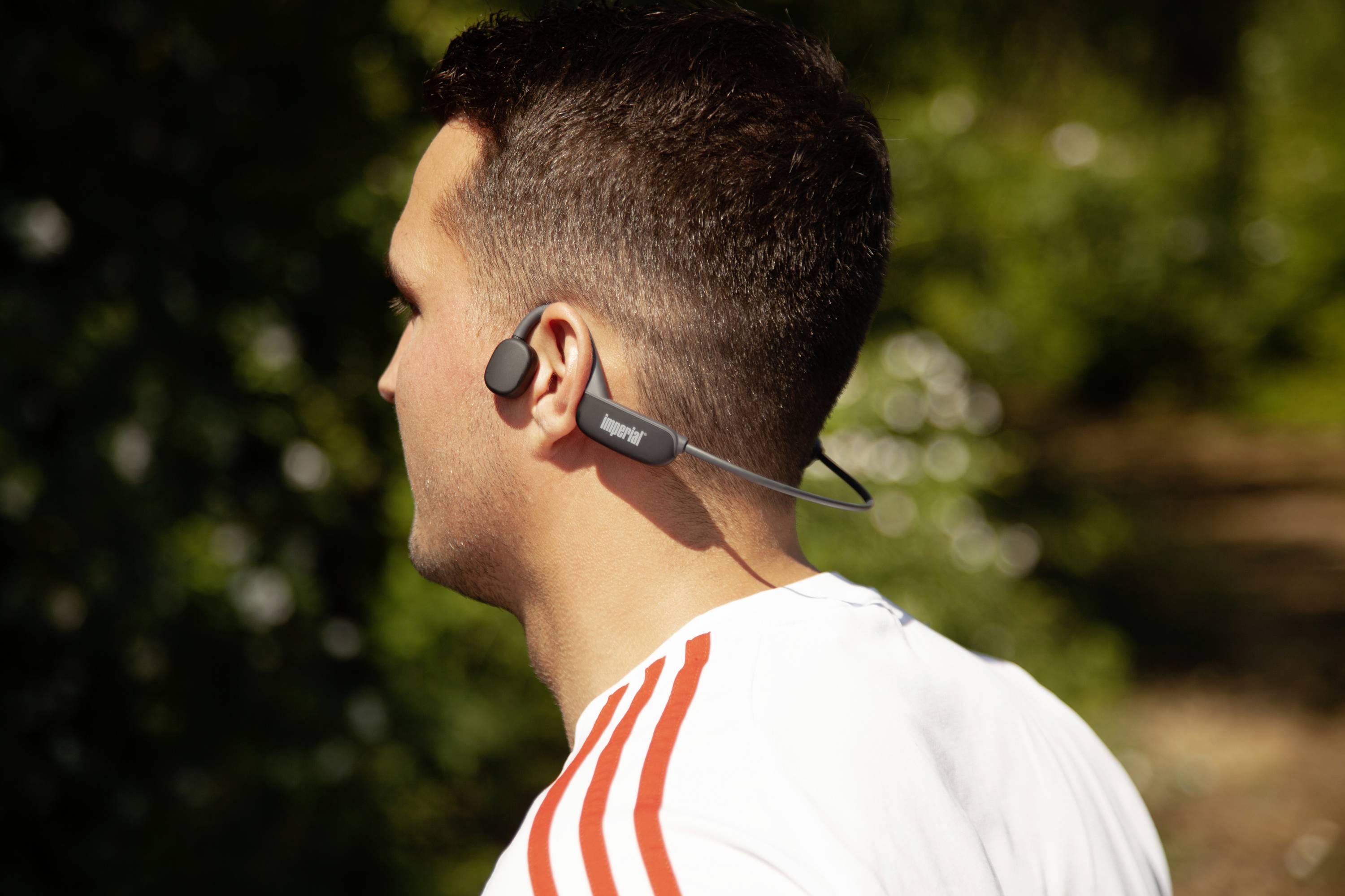 Une personne porte des écouteurs de sport sans fil à l'extérieur. La personne a les cheveux courts et porte un haut blanc avec des rayures rouges.