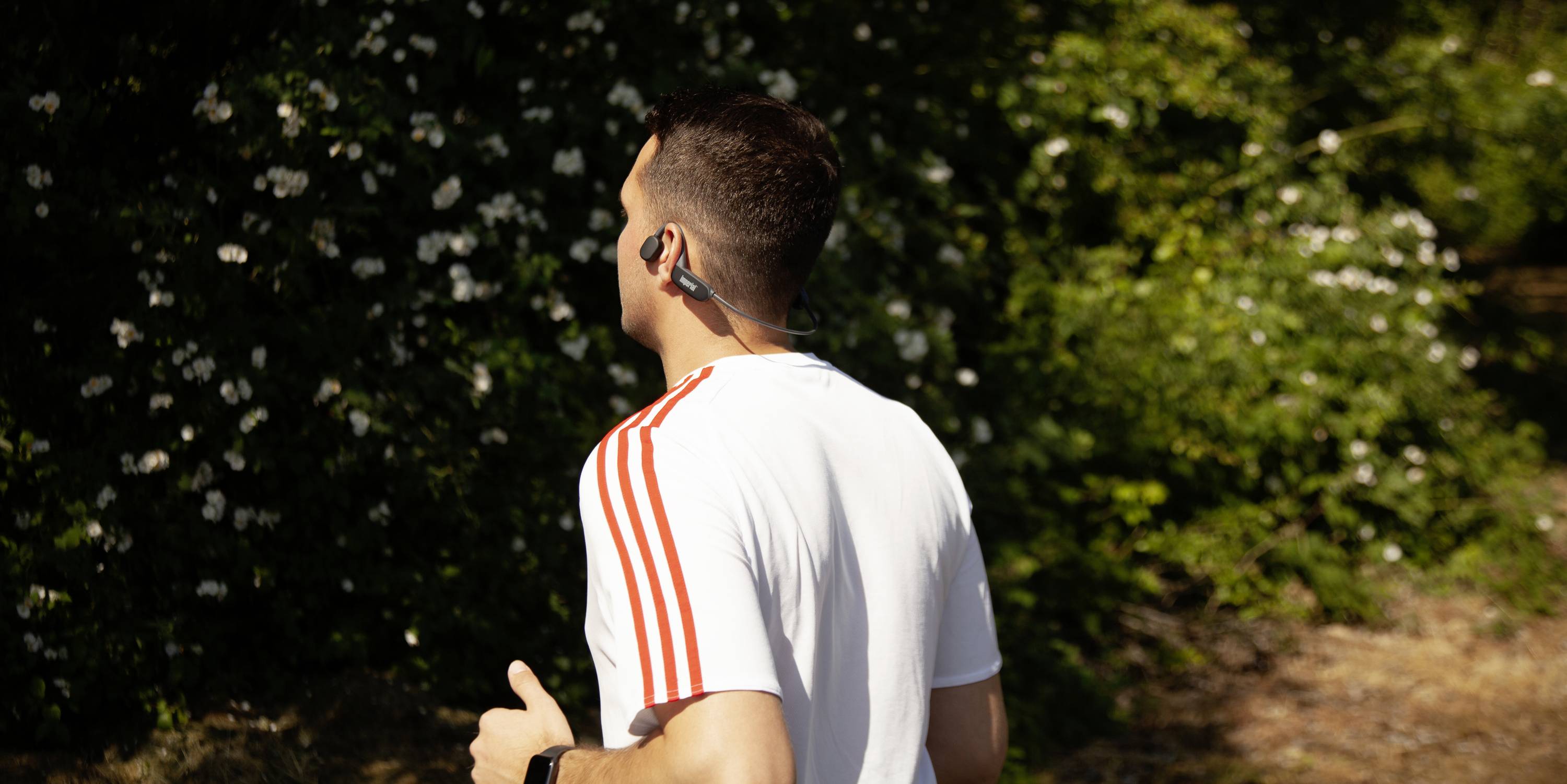 Un homme fait du jogging en extérieur, portant un t-shirt blanc avec des rayures rouges et des écouteurs. Des arbres et des arbustes verts sont visibles en arrière-plan.