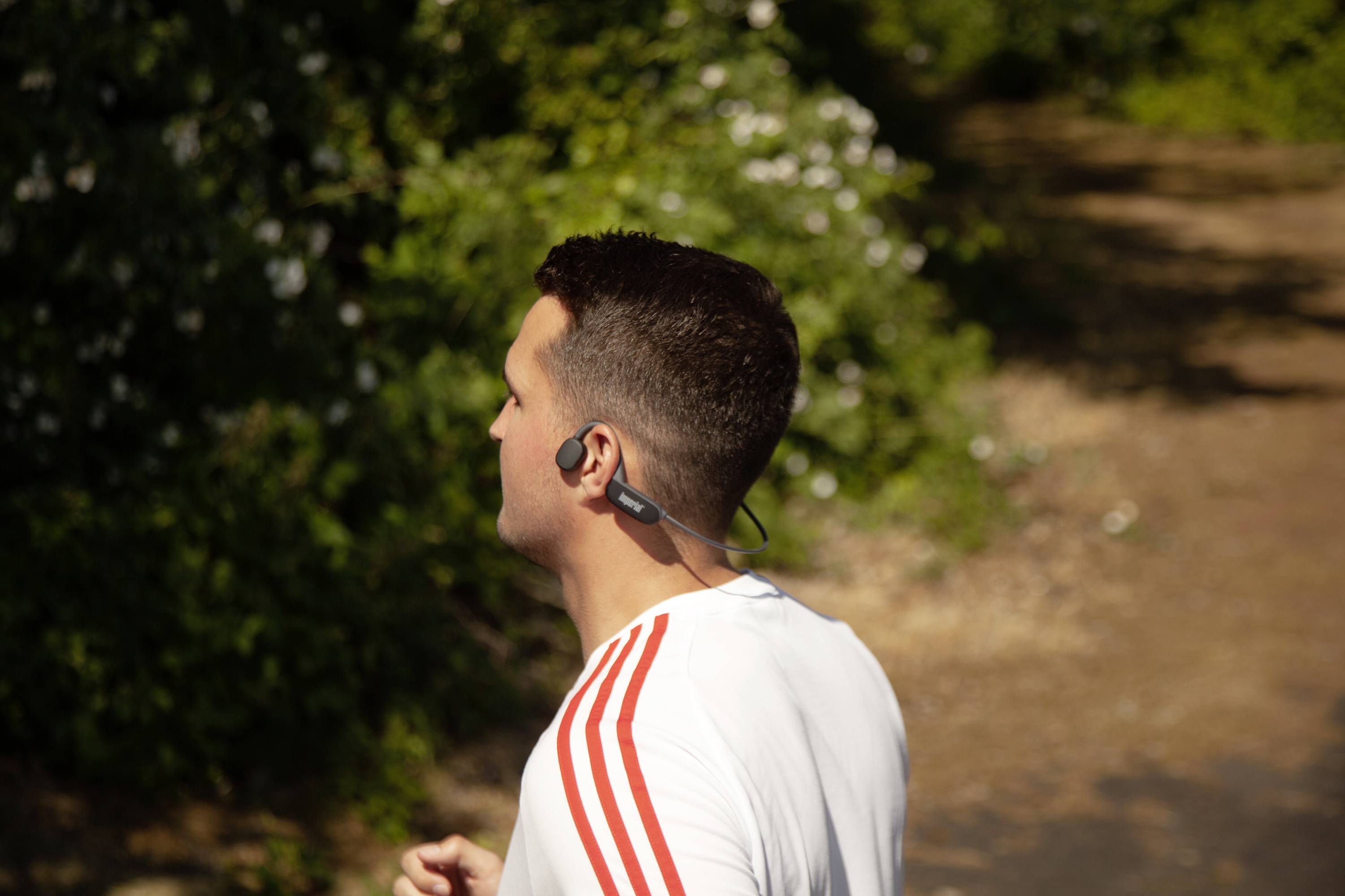 Un homme fait du jogging sur un chemin ensoleillé, portant un t-shirt blanc à rayures rouges et des écouteurs, entouré de végétation verdoyante.