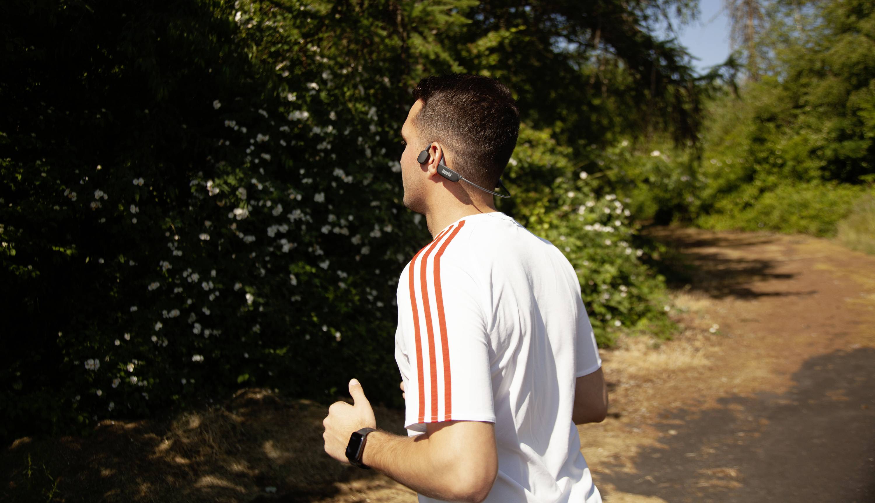 Une personne fait du jogging sur un sentier forestier ombragé, portant un t-shirt blanc à rayures rouges et des écouteurs aux oreilles.