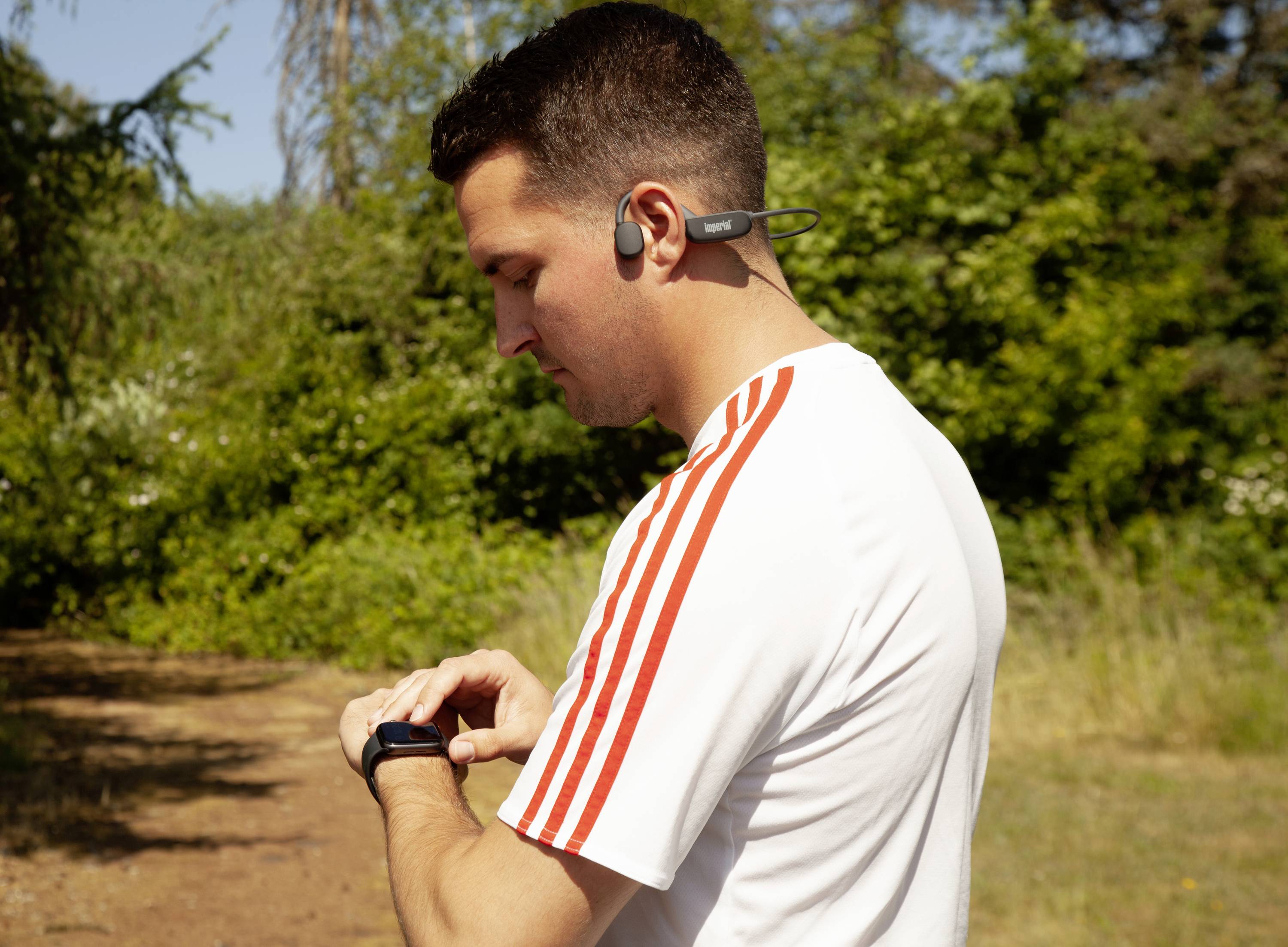 Un homme en tenue de sport portant un t-shirt blanc à rayures rouges regarde sa montre connectée. Il porte des écouteurs sans fil. En arrière-plan, des arbres verts.