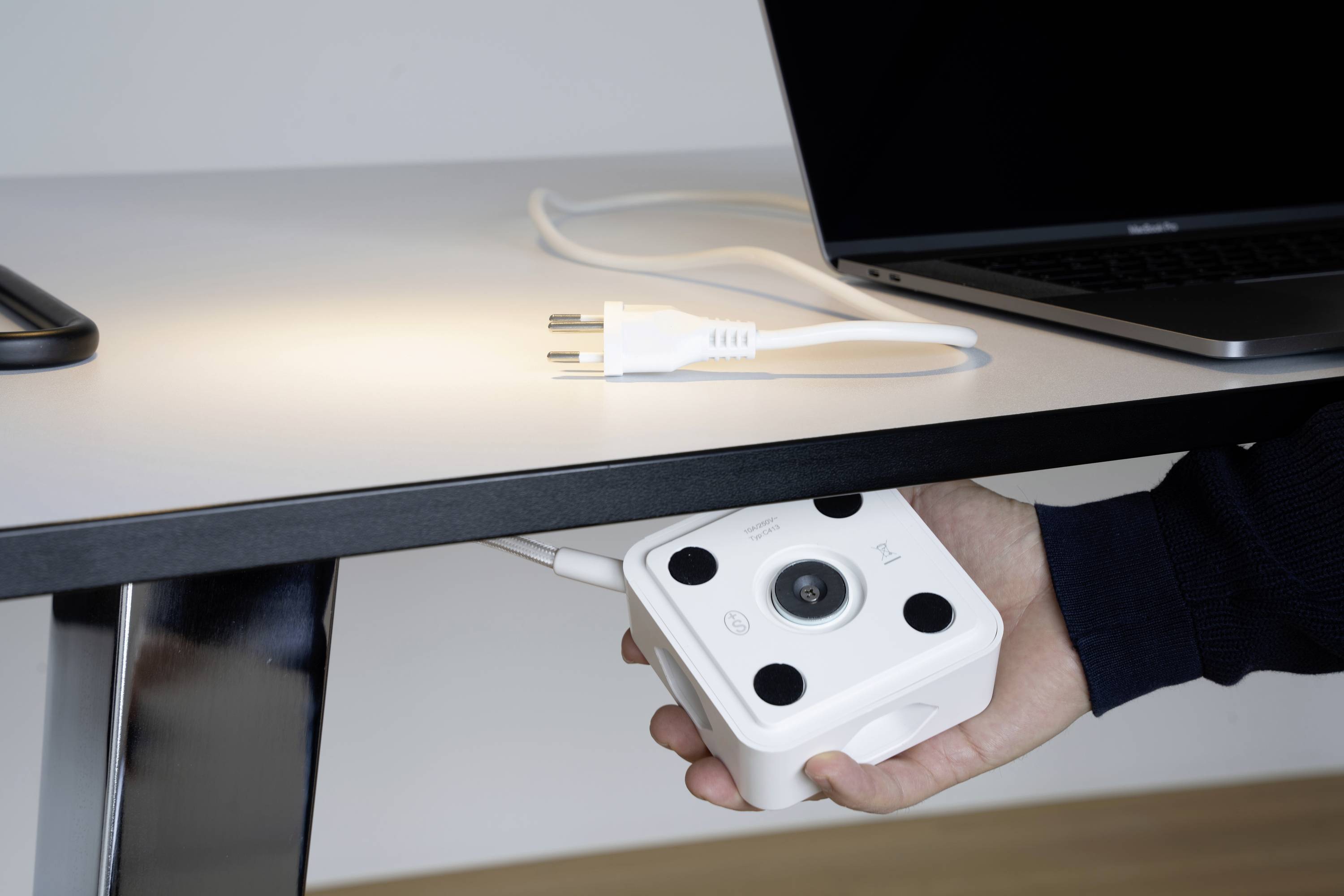 Une main tient un multiprise blanc sous une table. Sur la table se trouvent un ordinateur portable et une prise.