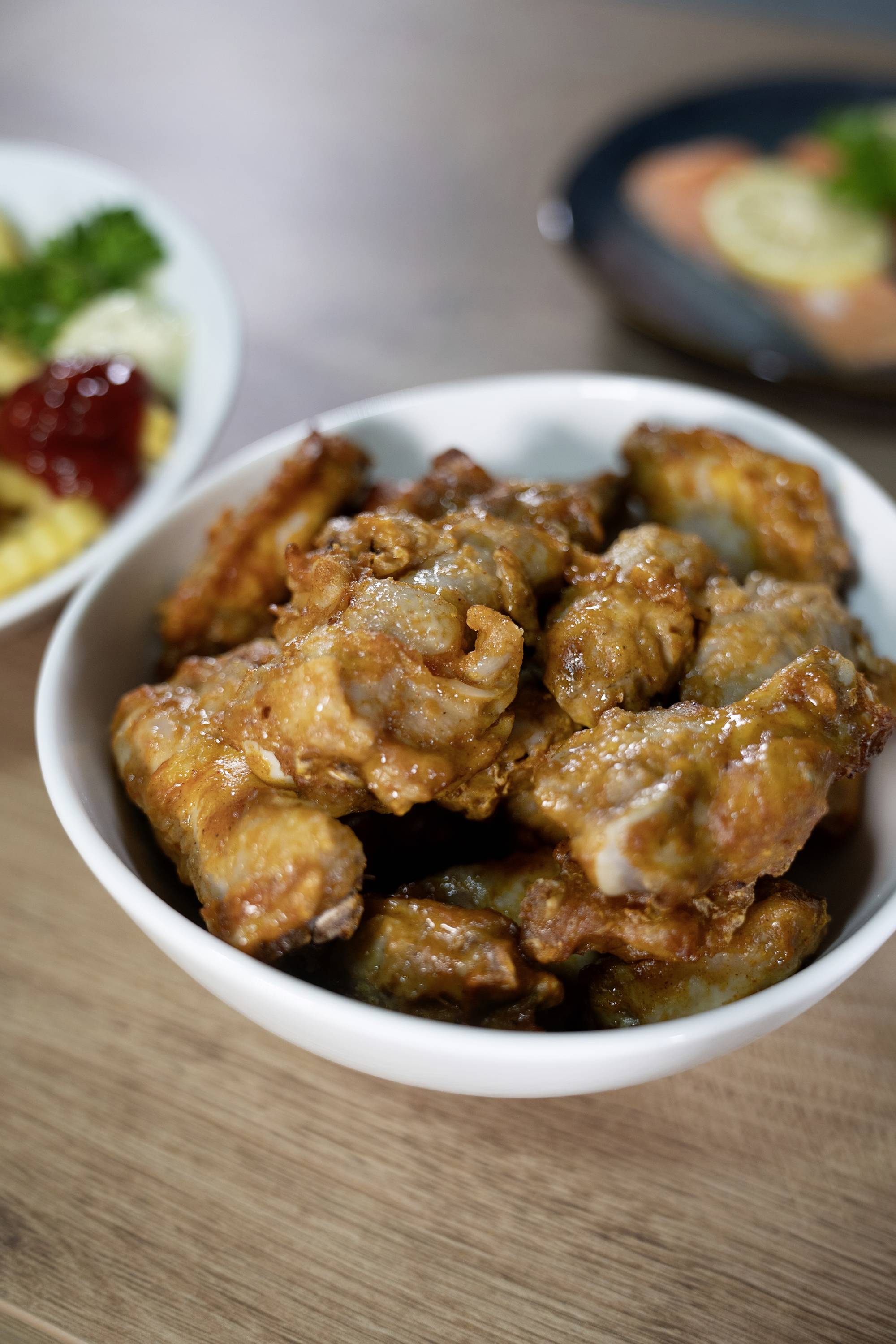Un bol de wings de poulet frits, placé à l'avant d'une table en bois, avec des accompagnements flous en arrière-plan.
