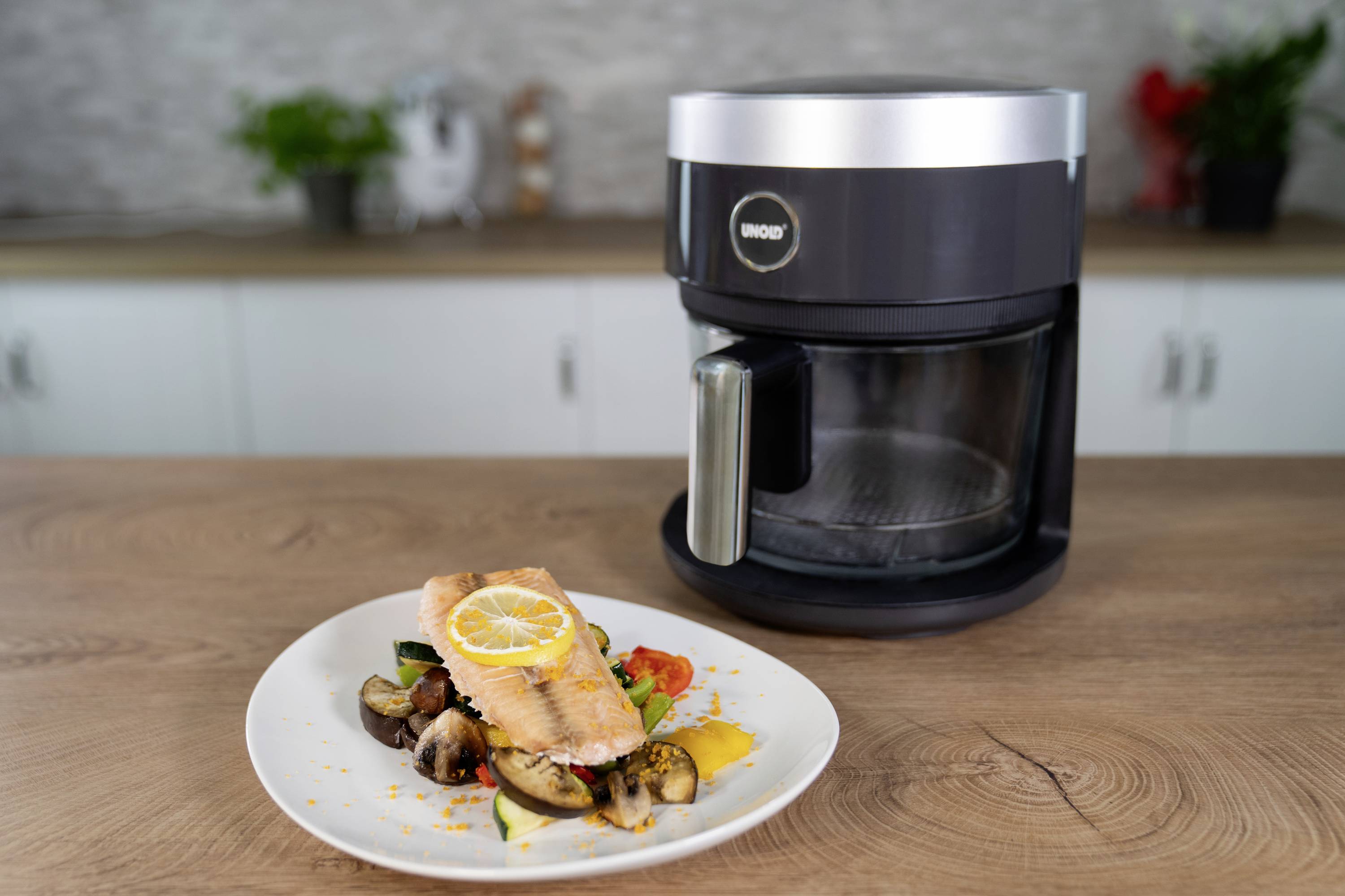 Un plat de saumon grillé sur des légumes devant un robot de cuisine noir sur un plan de travail en bois dans une cuisine moderne.