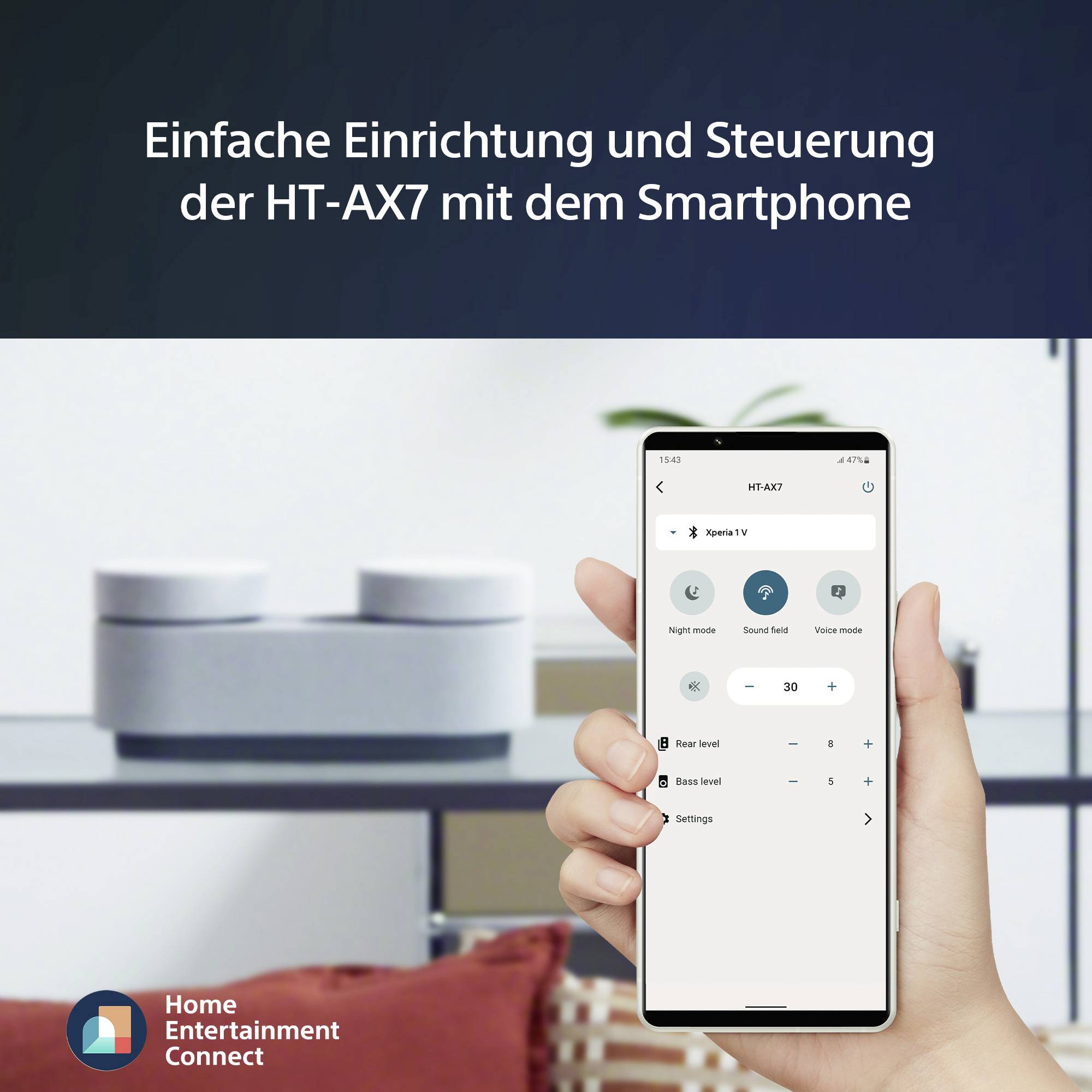 Un smartphone affiche l'application de contrôle de la HT-AX7. Un salon est discrètement visible en arrière-plan.