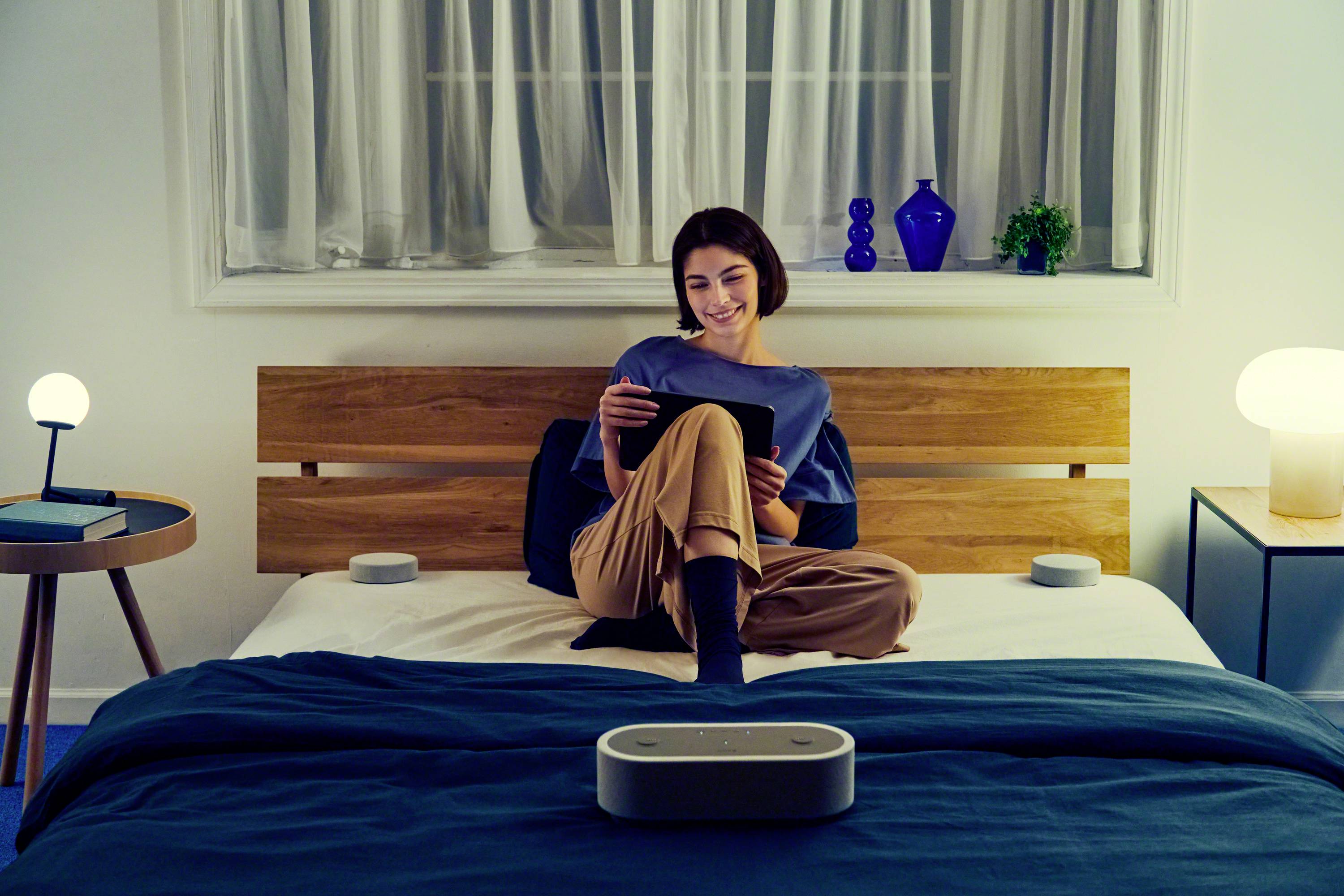 Une femme est assise sur un lit et lit quelque chose sur une tablette. Une petite lampe est posée sur une table à côté d'elle, et la pièce est doucement éclairée.