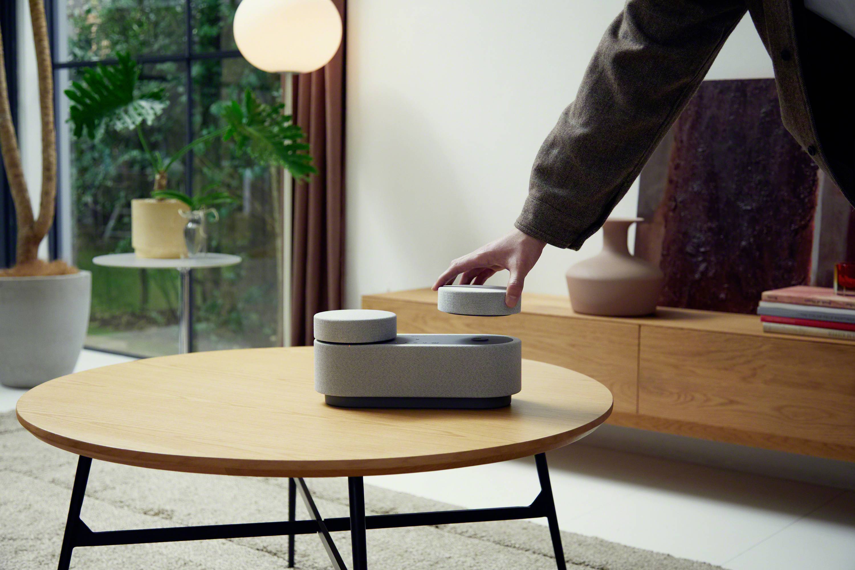 Une personne saisit un appareil rond gris et blanc sur une table en bois. En arrière-plan, des plantes et des meubles sont visibles dans une pièce moderne.