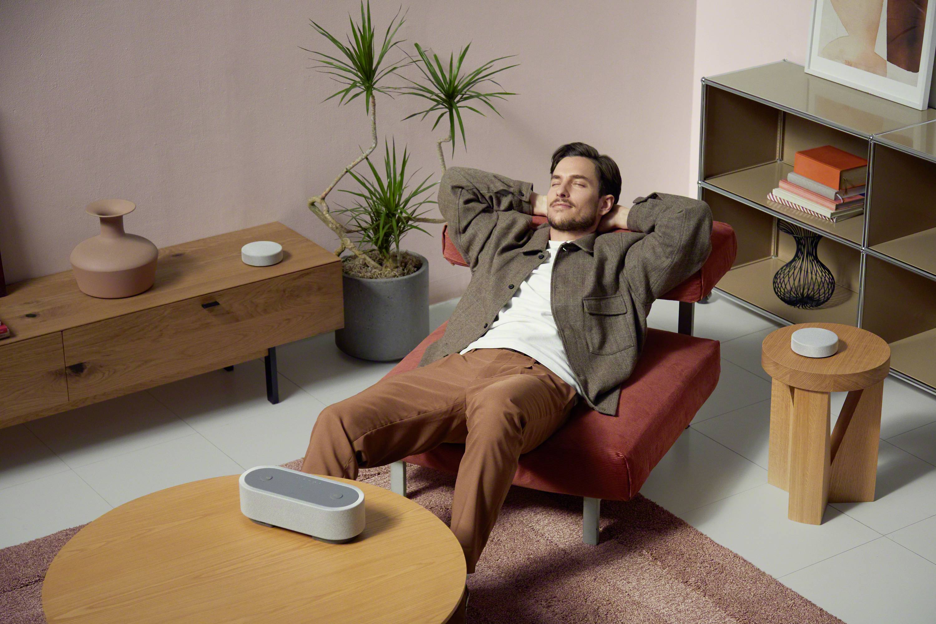 Un homme est allongé de manière détendue dans un fauteuil dans un salon au design moderne, avec des plantes et un haut-parleur sur la table.