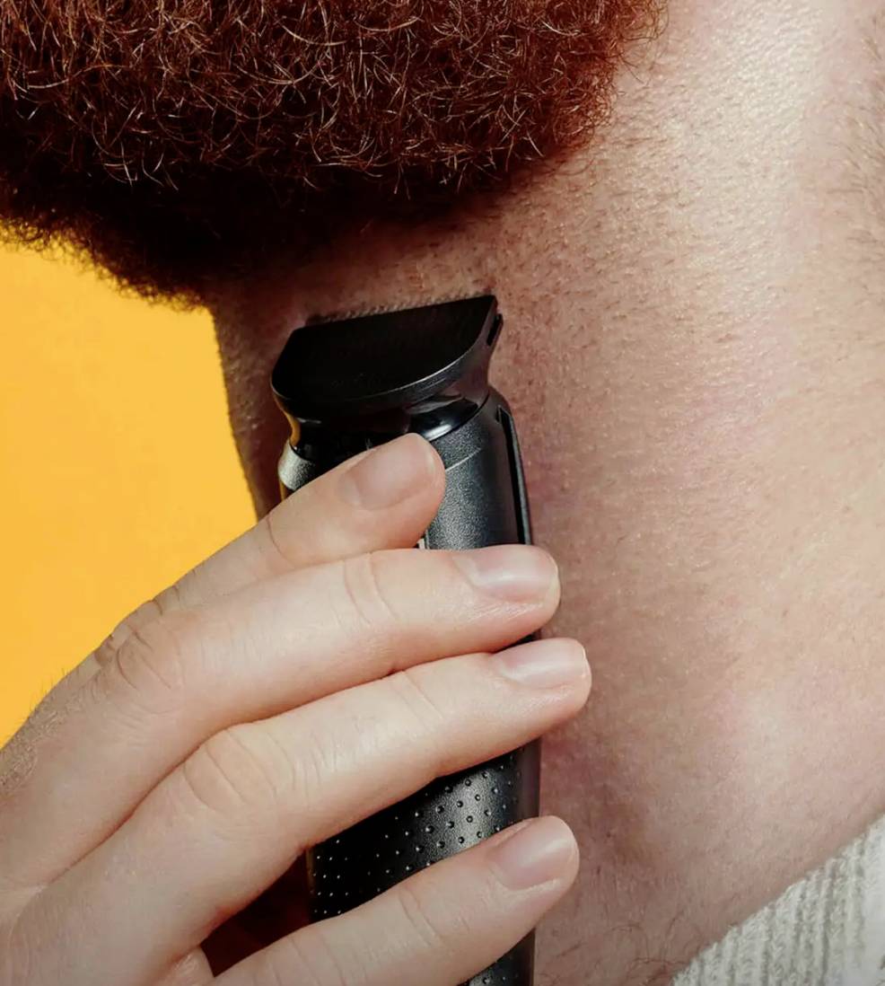 Une personne taille sa barbe avec un rasoir électrique au niveau de la ligne du cou. L'arrière-plan est orange, ce qui renforce la mise au point.