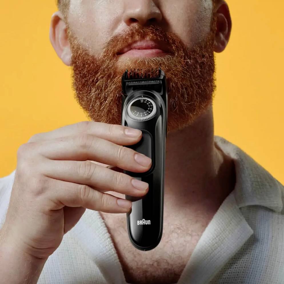 Un homme à la barbe rousse tient un rasoir électrique noir de Braun devant un fond orange.