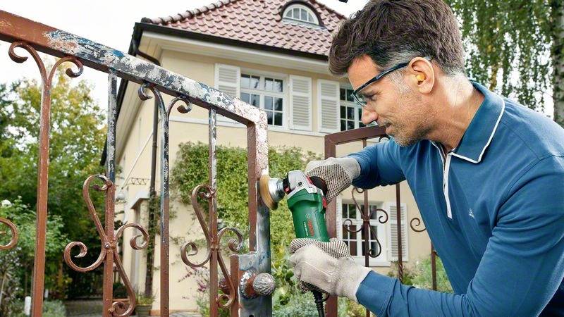 Un homme prépare un portail métallique à l'aide d'une meuleuse devant une grande maison.