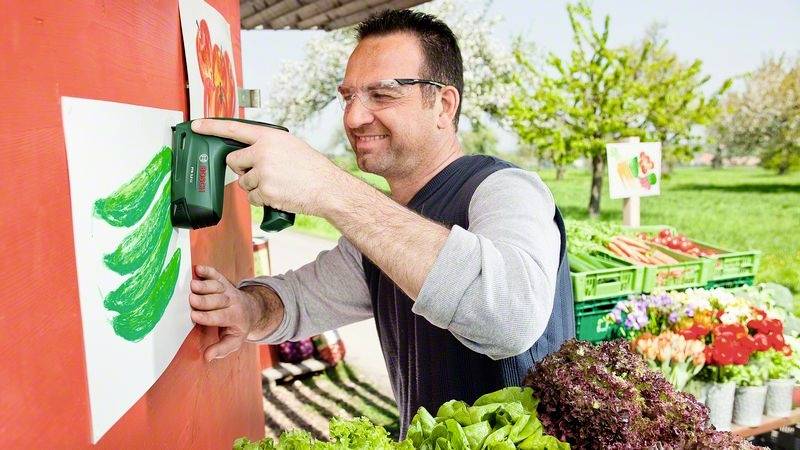 Un homme fixe une affiche avec des concombres verts sur un mur rouge, entouré de tables remplies de légumes frais sur un marché en plein air.