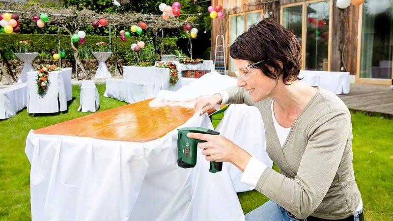Une femme nettoie une table à l'extérieur en vue d'un événement. La table est recouverte d'un tissu blanc. Des ballons sont visibles en arrière-plan.
