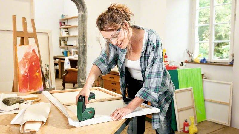 Une femme dans un atelier tend une toile sur un cadre en bois à l'aide d'un outil électrique. Des accessoires de peinture sont visibles en arrière-plan.