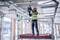 Un ouvrier portant un gilet de sécurité et un casque de chantier est debout sur un échafaudage et effectue l'installation du plafond dans un bâtiment.