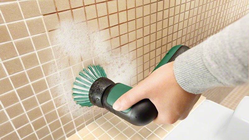 Une personne nettoie des carreaux beiges à l'aide d'une brosse électrique, en appliquant un spray nettoyant.