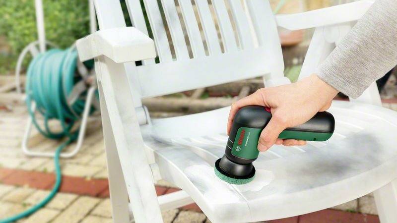 Une personne nettoie une chaise blanche en plastique à l'aide d'un petit appareil de nettoyage portatif. Un tuyau d'arrosage est visible en arrière-plan.