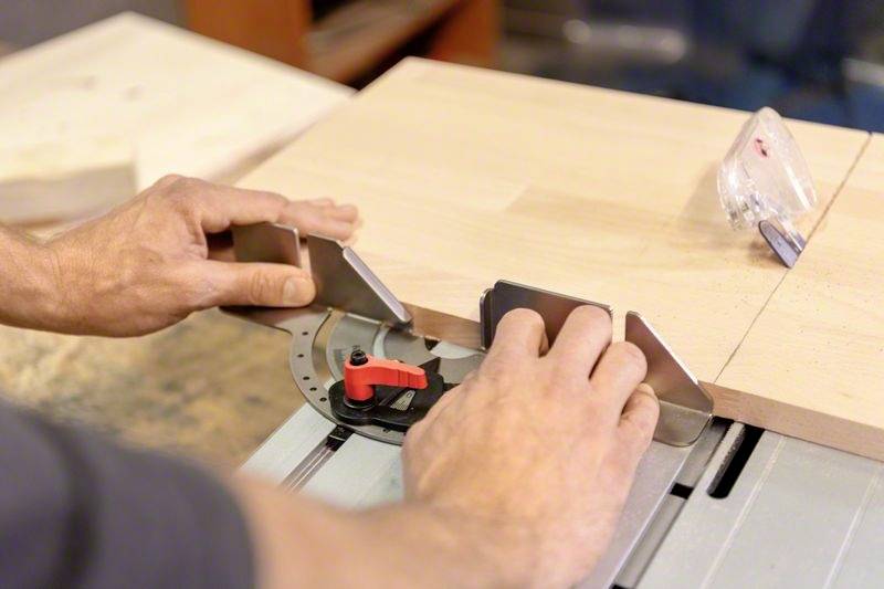 Deux mains ajustent une planche en bois sur une scie circulaire de table avec une butée d'angle. Préparation pour une coupe précise.