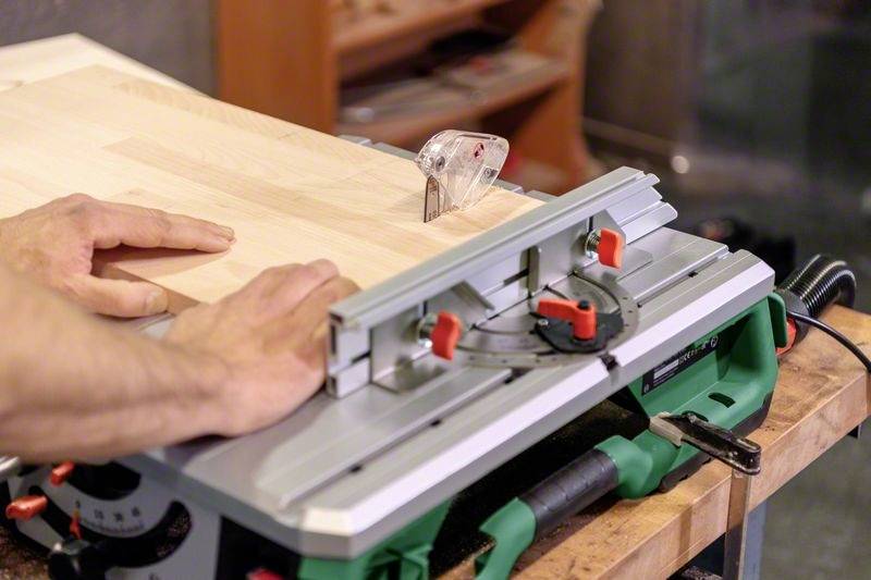 Une personne utilise une scie circulaire de table verte pour couper un morceau de bois. Les mains guident soigneusement le bois.