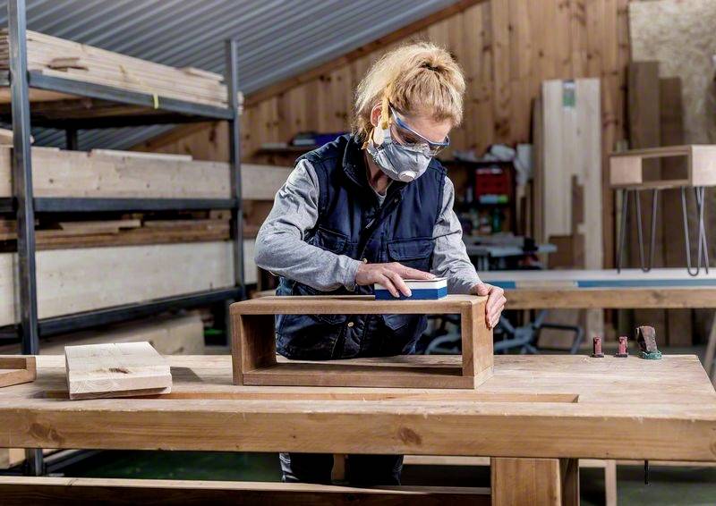 Une personne travaille dans un atelier de menuiserie, ponçant une pièce de bois sur une grande table. Des étagères avec des matériaux en bois sont visibles en arrière-plan.