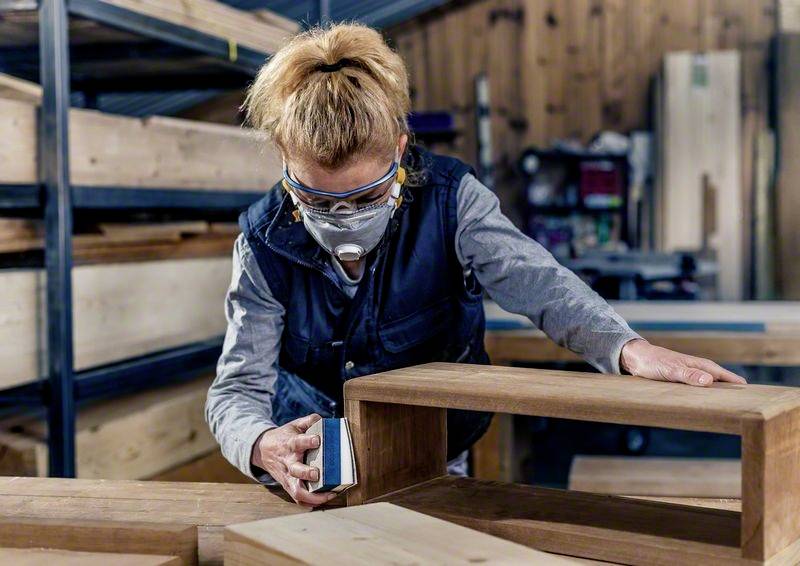 Une personne travaille un morceau de bois dans un atelier. Elle porte des lunettes de protection et un masque, tandis qu'elle ponce soigneusement le bois. Des étagères en bois sont visibles en arrière-plan.