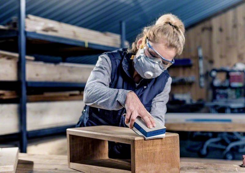Une personne ponce un morceau de bois dans un atelier. Elle porte des lunettes de protection et un masque respiratoire. L'arrière-plan montre des étagères en bois et des outils.