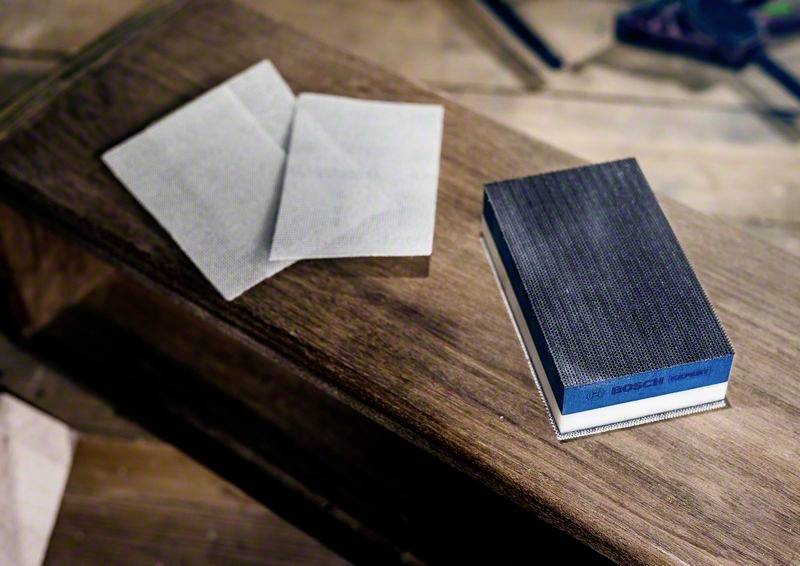 Un bloc de ponçage et deux feuilles de papier abrasif sont posés sur une table en bois. Des outils sont visibles en arrière-plan.