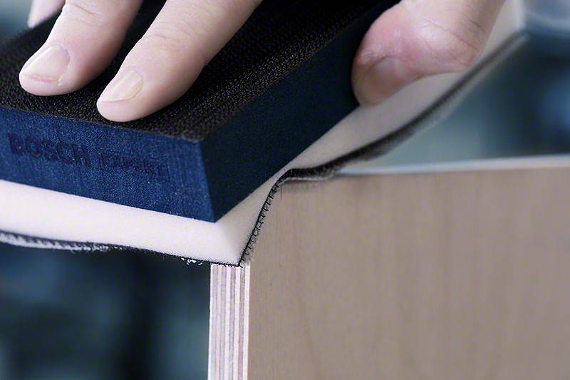 Une main utilise une éponge à poncer pour lisser le bord d'une planche en bois, ce qui suggère une activité artisanale.