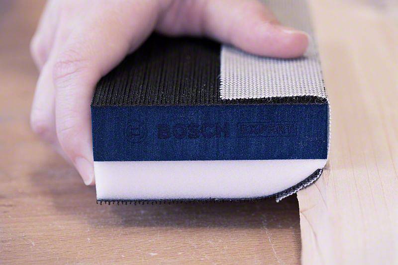'Une main tient une éponge de ponçage de la marque Bosch sur une planche en bois, prête à poncer.'