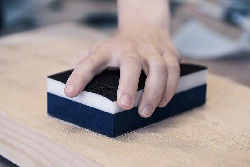 Une personne tient un bloc de ponçage rectangulaire sur une planche de bois, probablement en train de poncer une surface.
