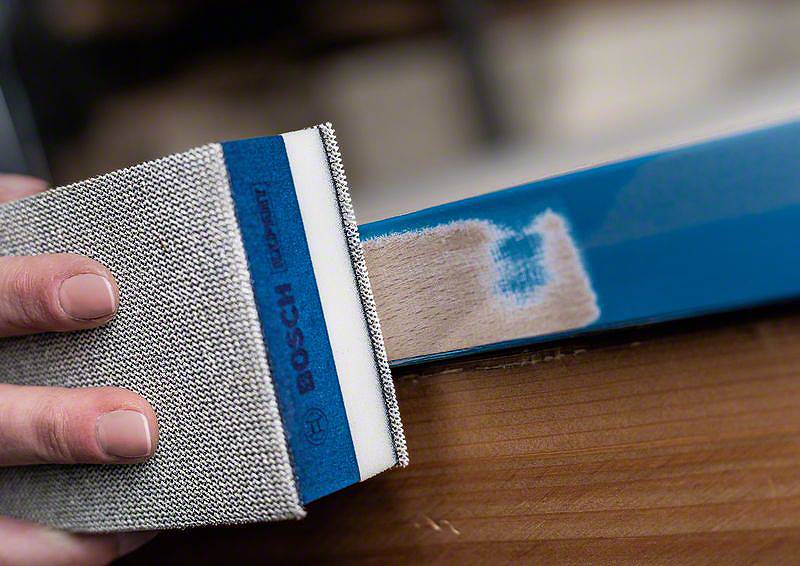 Une main ponce le bord bleu d'une pièce en bois avec un bloc de ponçage. La couleur du bois devient visible. L'accent est mis sur la précision et l'artisanat.