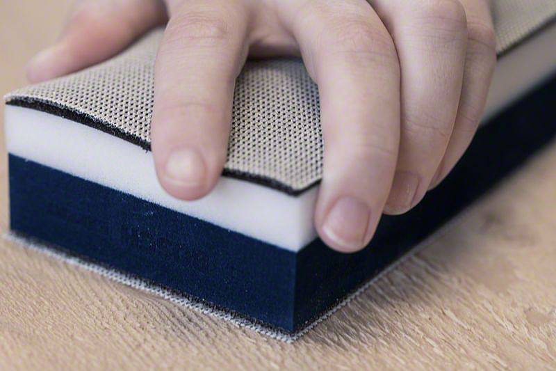 Une main tient une éponge rectangulaire aux couches blanches, grises et bleues sur une surface en bois.