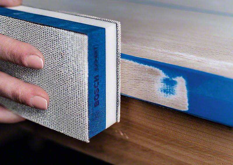 Une main ponce le bord d'une table avec un bloc de ponçage bleu pour lisser la surface.