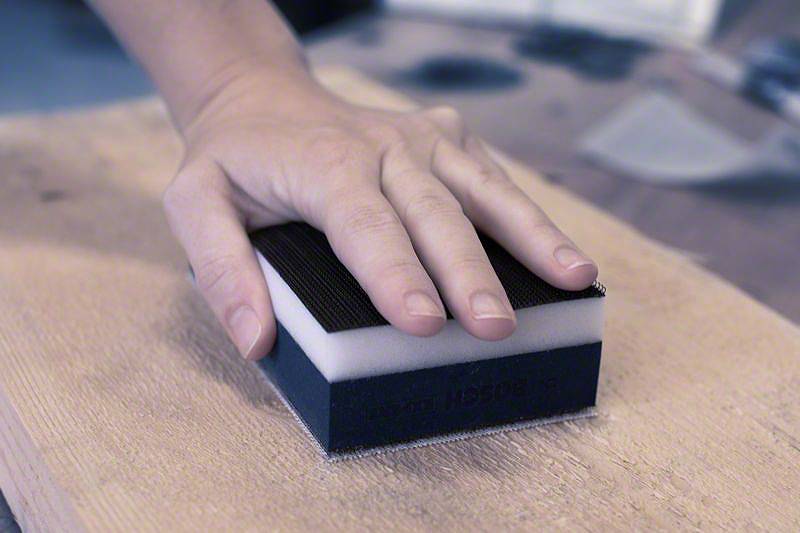 Une main tient une éponge abrasive et travaille une surface en bois. L'éponge a un côté noir de ponçage.
