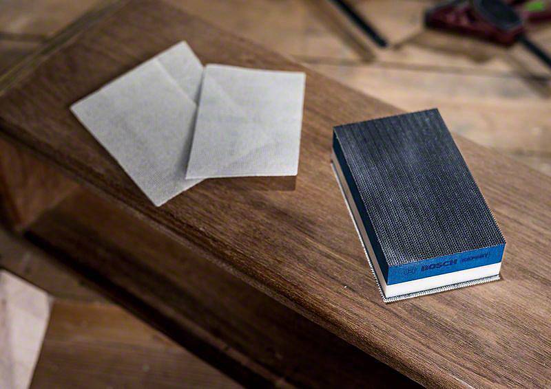 Un bloc de ponçage et deux feuilles de papier de verre sont posés sur une table en bois, prêts à être utilisés pour le travail du bois.