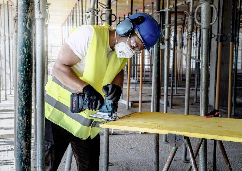 Ouvrier portant des équipements de protection et des protections auditives en train de couper du bois sur un chantier de construction.