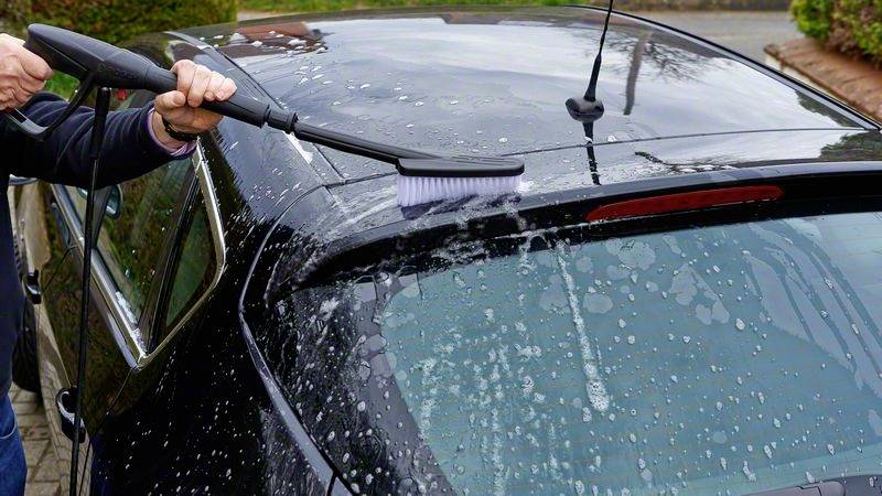 Une personne nettoie le toit d'une voiture noire avec une brosse et de l'eau savonneuse. La voiture est garée à l'extérieur sur une surface pavée.
