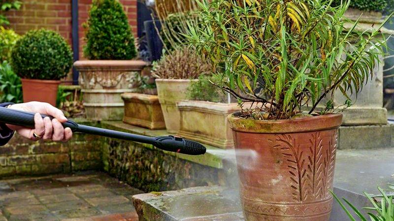 Une personne nettoie un pot de fleurs en terre cuite avec un nettoyeur haute pression dans un jardin, entourée de plusieurs plantes.