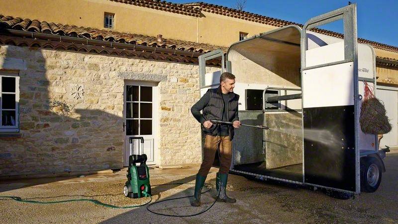 Un homme nettoie l'intérieur d'un van pour chevaux à l'aide d'un nettoyeur haute pression devant un bâtiment en pierre.