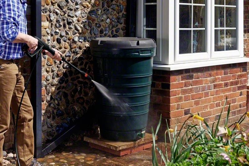 Une personne utilise un nettoyeur haute pression pour nettoyer un grand récipient de collecte d'eau de pluie vert à côté d'une maison en briques.
