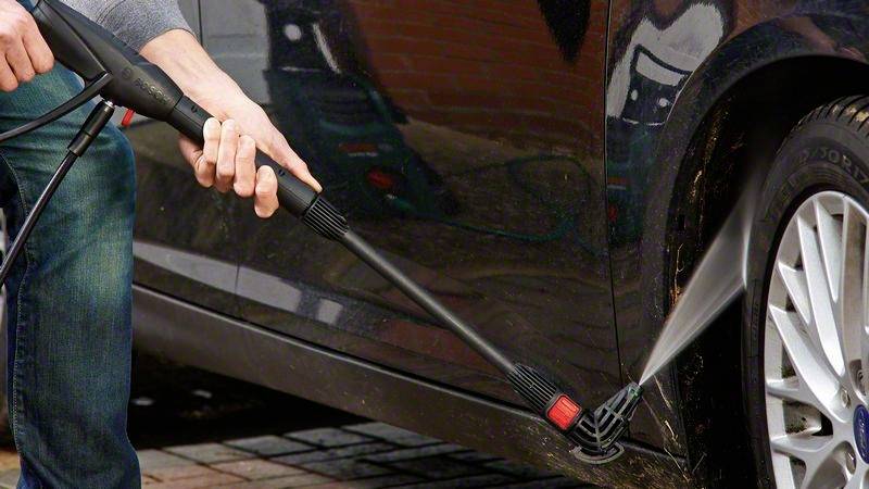 Une personne nettoie la partie inférieure d'une voiture avec un nettoyeur haute pression, pulvérisant de l'eau sur la zone des roues pour enlever la saleté.