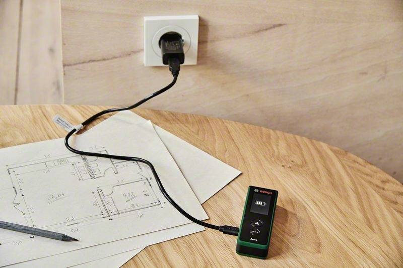 Un télémètre laser vert est posé sur une table en bois et mesure un dessin technique. Un câble est branché dans une prise électrique.