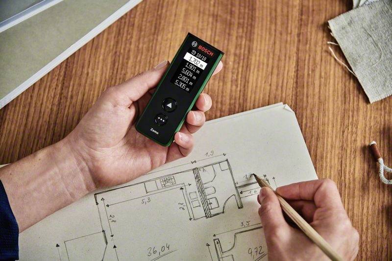 Une personne mesure des dimensions sur un plan technique pour un espace, à l'aide d'un télémètre numérique, posé sur une table en bois.