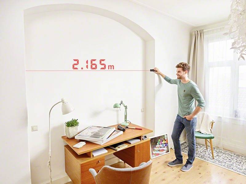 Une personne mesure la distance d'un mur dans une pièce lumineuse à l'aide d'un télémètre laser ; l'affichage indique '2,165 m'.