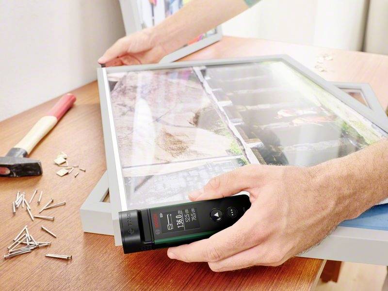 Une personne fixe un cadre photo sur le mur à l'aide d'un appareil de mesure numérique pour l'alignement. Des outils et des clous sont posés sur la table.