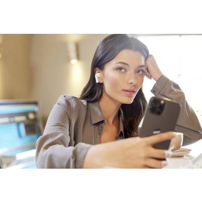 Une personne est assise à une table, tenant un smartphone et portant des écouteurs sans fil. Un écran d'ordinateur est visible en arrière-plan.