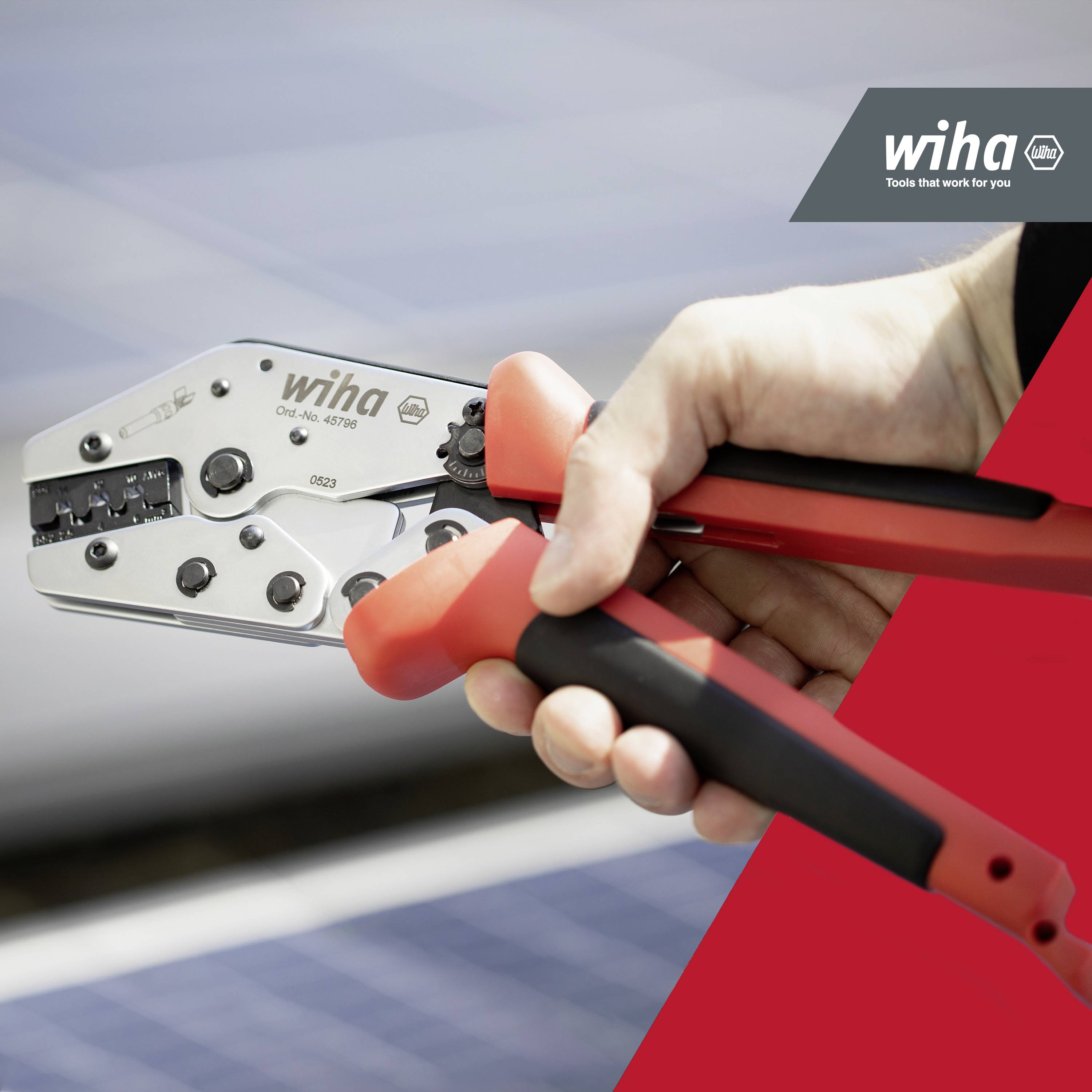 Une main tient une pince à sertir rouge et noire de la marque Wiha devant un arrière-plan flou. Le logo Wiha se trouve en haut à droite.